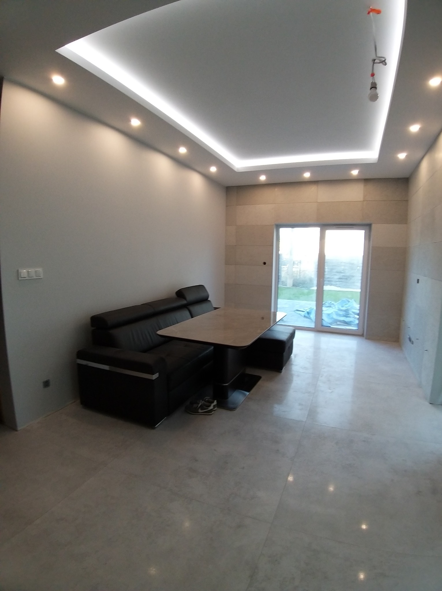 Wykończone wnętrze salonu z szarą podłogą, ścianami i sufitem z oświetleniem LED. Widoczny czarny narożnik i stół. Przesuwne drzwi balkonowe w tle.