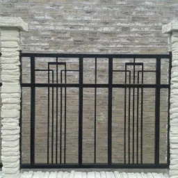 Fragment czarnego, metalowego ogrodzenia z geometrycznym wzorem, wspartego na dwóch kamiennych, teksturowanych filarach na tle ceglanego muru.