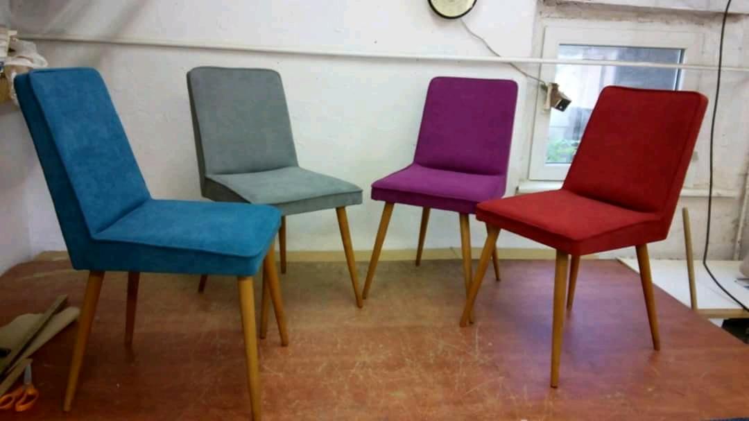 Cztery krzesła po renowacji tapicerskiej w Gdańsku: turkusowe, szare, fioletowe i czerwone, każde z drewnianymi nogami, ustawione na stole w warsztacie.