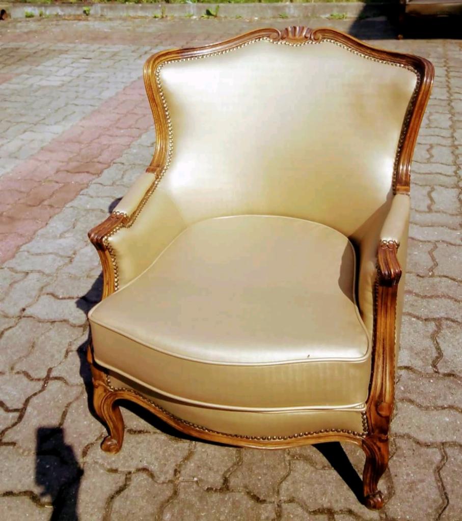 Odnowiony, beżowy fotel w stylu Ludwika Filipa z ciemnobrązową, rzeźbioną ramą i ozdobnymi gwoździami, sfotografowany na brukowanej nawierzchni.