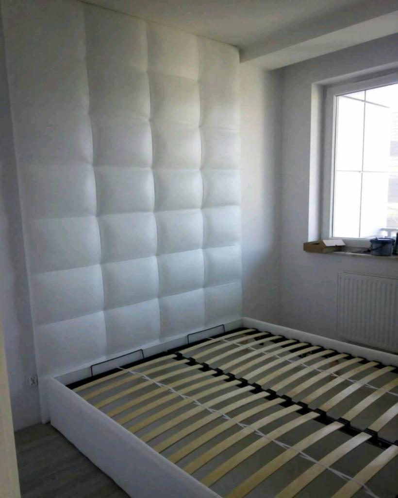 Białe, tapicerowane wezgłowie łóżka z kwadratowymi panelami, widok na drewniany stelaż w ramie łóżka, jasne wnętrze sypialni z oknem.