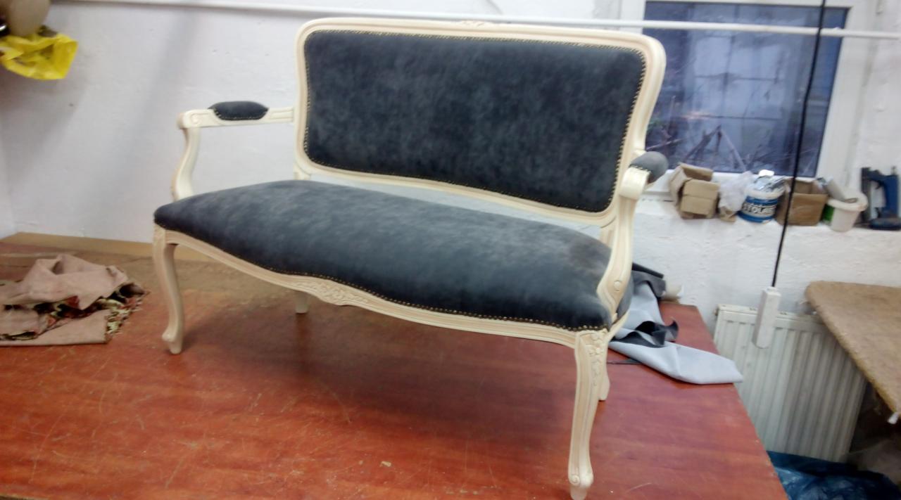 Odrestaurowana kanapa w stylu Ludwika, z szarym obiciem i kremowymi zdobieniami, na warsztatowym stole.