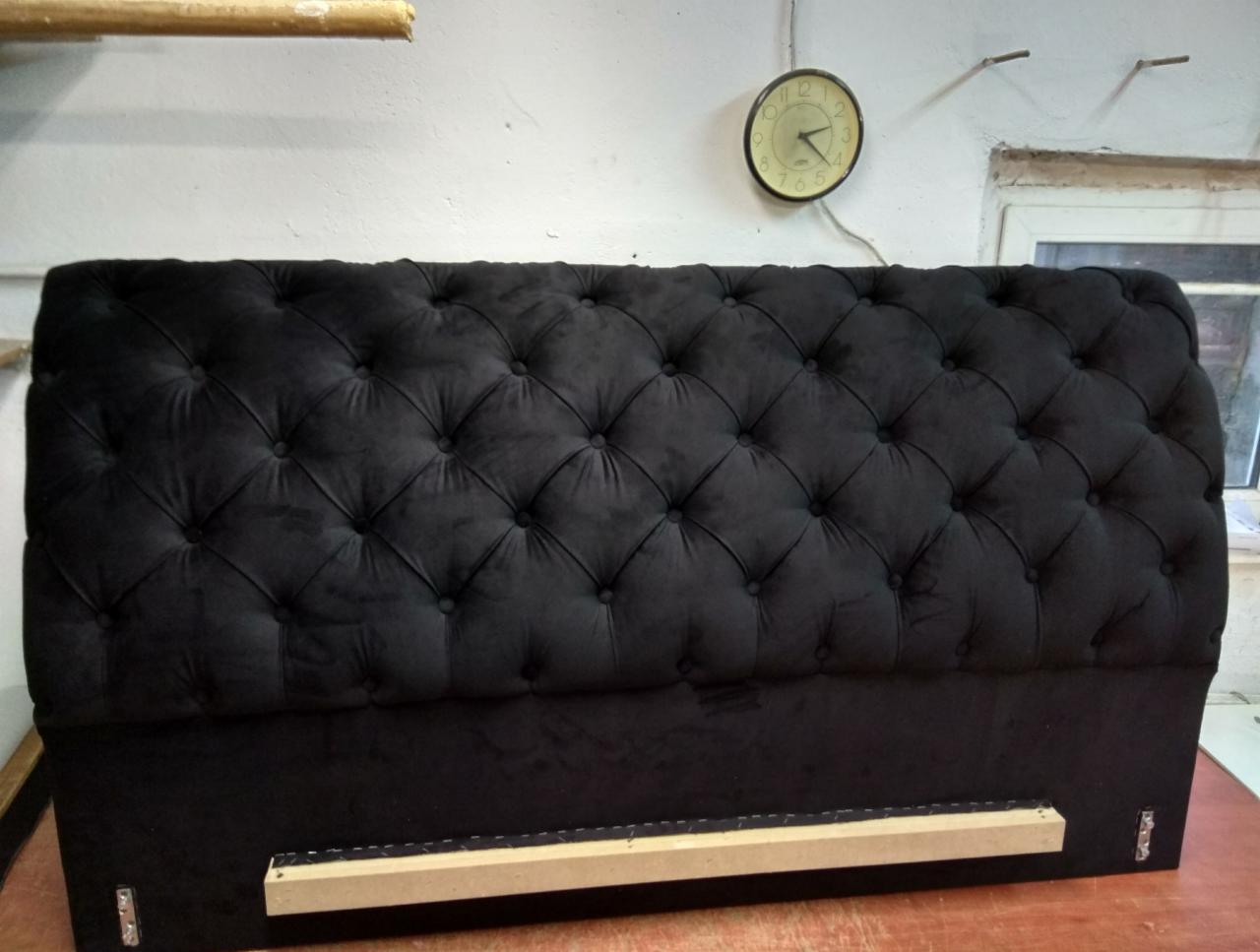 Tapicerowane, czarne wezgłowie łóżka w stylu chesterfield z widocznymi guzikami i drewnianą konstrukcją mocującą, sfotografowane w warsztacie, z zegarem na ścianie w tle.