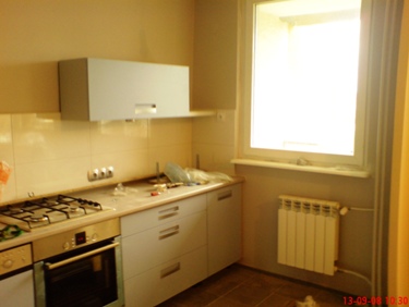 Kuchnia w trakcie remontu: zamontowane jasnoniebieskie szafki wiszące i dolne, widoczne narzędzia na blacie, grzejnik pod oknem, płytki na ścianie i częściowo zabezpieczona folią kuchenka.