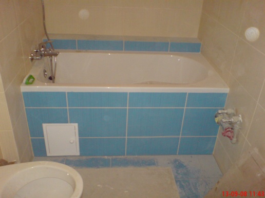 Wykończona łazienka z wanną obłożoną błękitnymi płytkami, widoczna armatura, biała klapa rewizyjna i fragment toalety w dolnym rogu kadru.