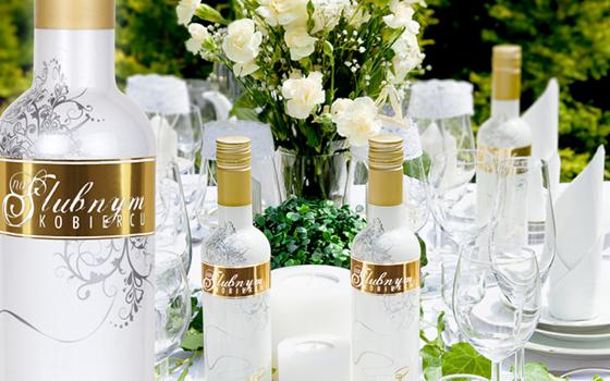 Dekoracyjna butelka z etykietą 'Na Ślubnym Kobiercu' na stole weselnym z białymi kwiatami i szkłem.
