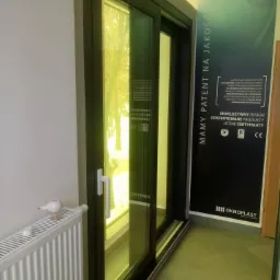 Czarna, przesuwna brama okienna z białym uchwytem, widoczny grzejnik i reklama firmy Oknoplast z hasłem 'Mamy patent na jakość'.