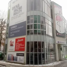 Budynek biurowy z dużą ilością przeszkleń i reklamami firm oferujących stolarkę okienną i drzwiową, widoczne reklamy Oknoplast, Wiśniowski oraz inne firmy z branży motoryzacyjnej.