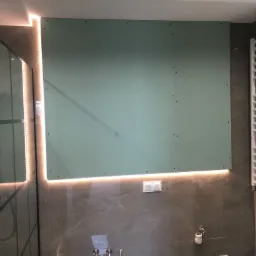 Ściana w łazience z zamontowaną płytą gipsowo-kartonową, podświetleniem LED i widocznymi otworami po wkrętach, obok kabina prysznicowa i grzejnik drabinkowy.