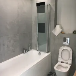 Nowoczesna łazienka po remoncie: biała wanna z parawanem nawannowym, podwieszana toaleta z otwartą klapą, szare płytki imitujące beton na ścianach, wzorzysta podłoga, rolka papieru i banan na półce.