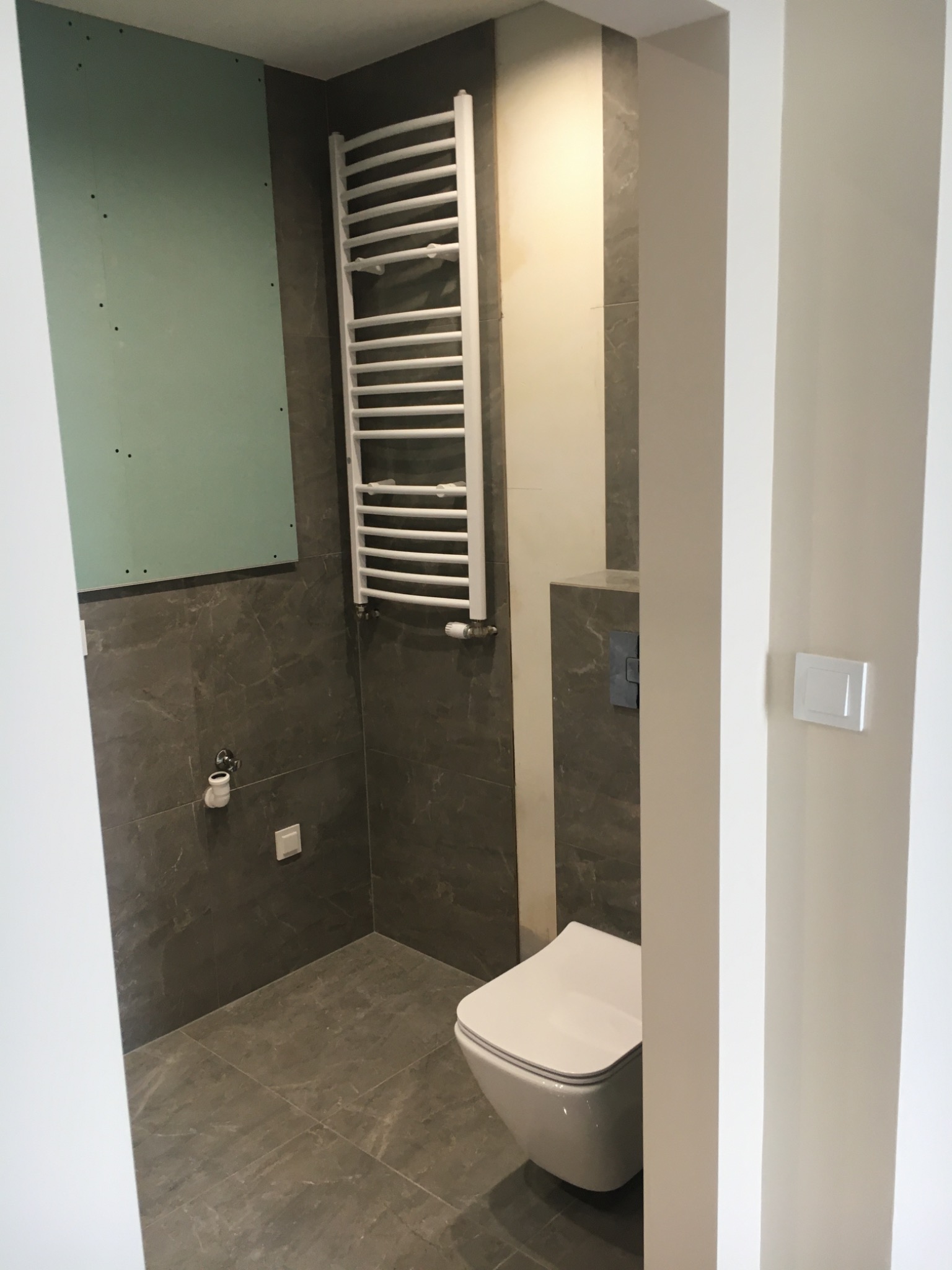 Wnętrze łazienki w trakcie remontu: ściana z płytą kartonowo-gipsową, grzejnik drabinkowy, podwieszana toaleta z przyciskiem spłukującym, szara płytka imitująca kamień na ścianach i podłodze, widok...