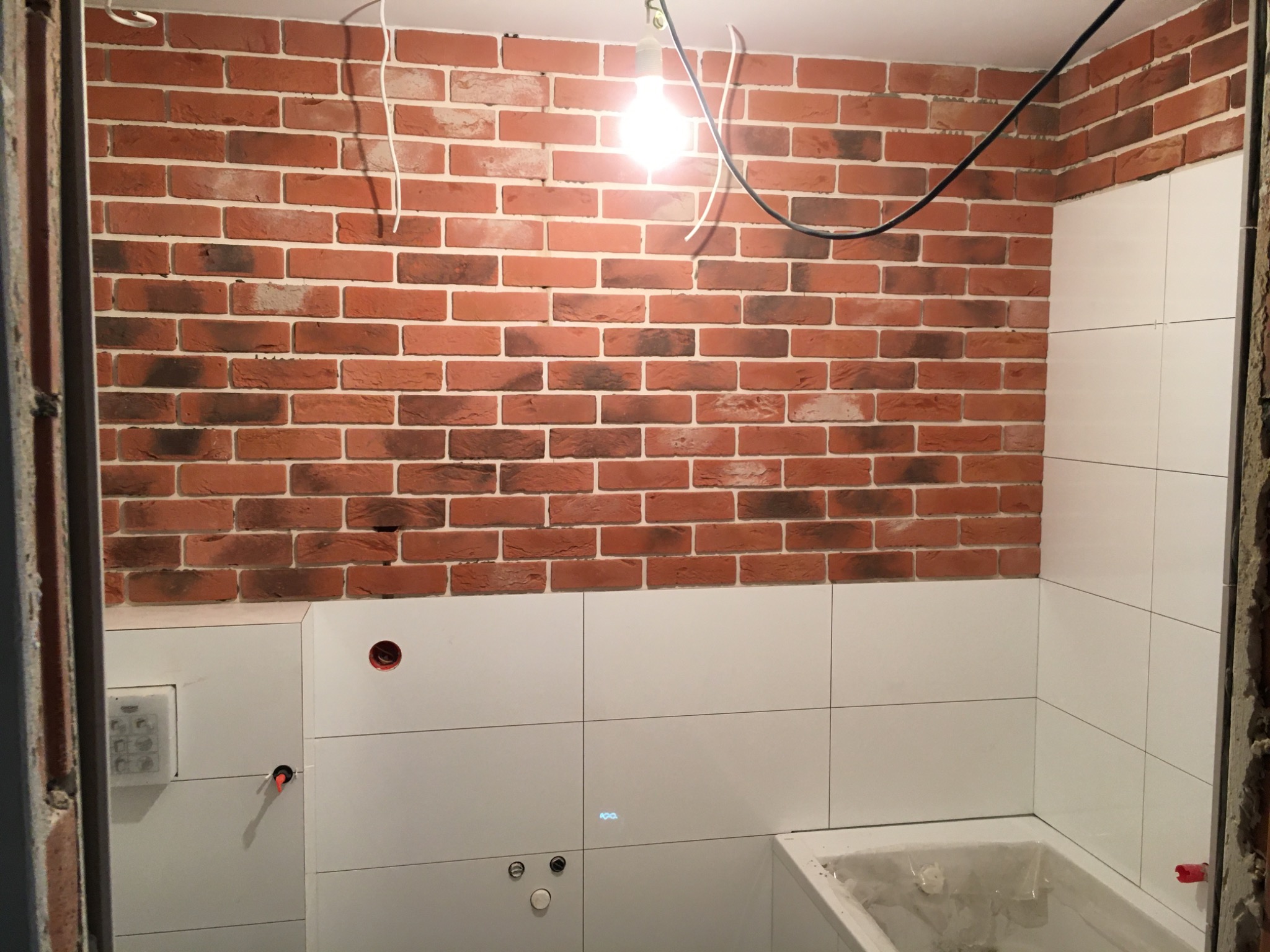 Ściana w łazience wykończona czerwoną cegłą i białymi płytkami, widoczne wystające przewody elektryczne i niedokończone elementy instalacji wodnej.