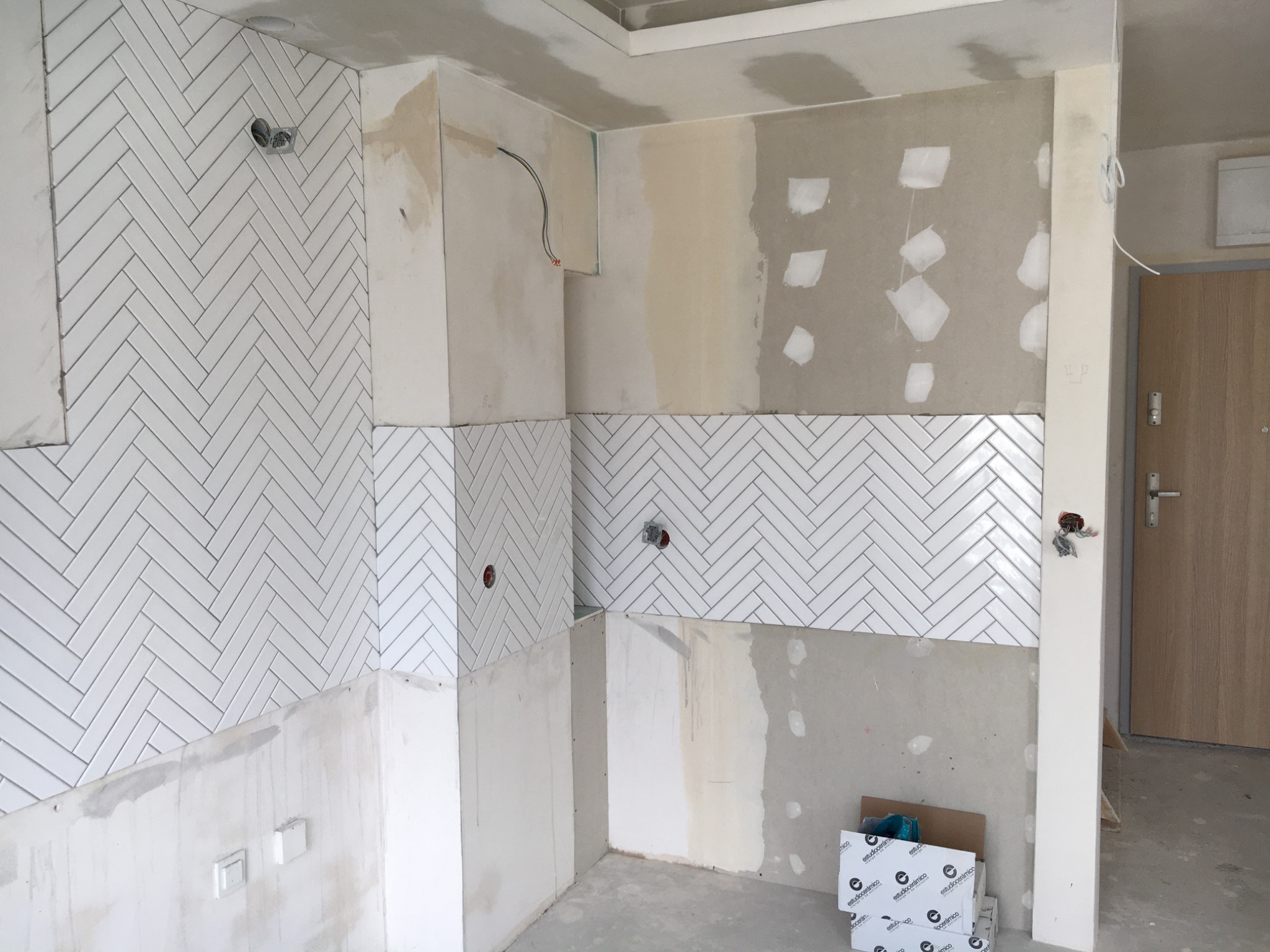 Montaż białych płytek w jodełkę na ścianie w trakcie remontu kuchni, widoczne puszki elektryczne, surowy tynk i kartony z płytkami.