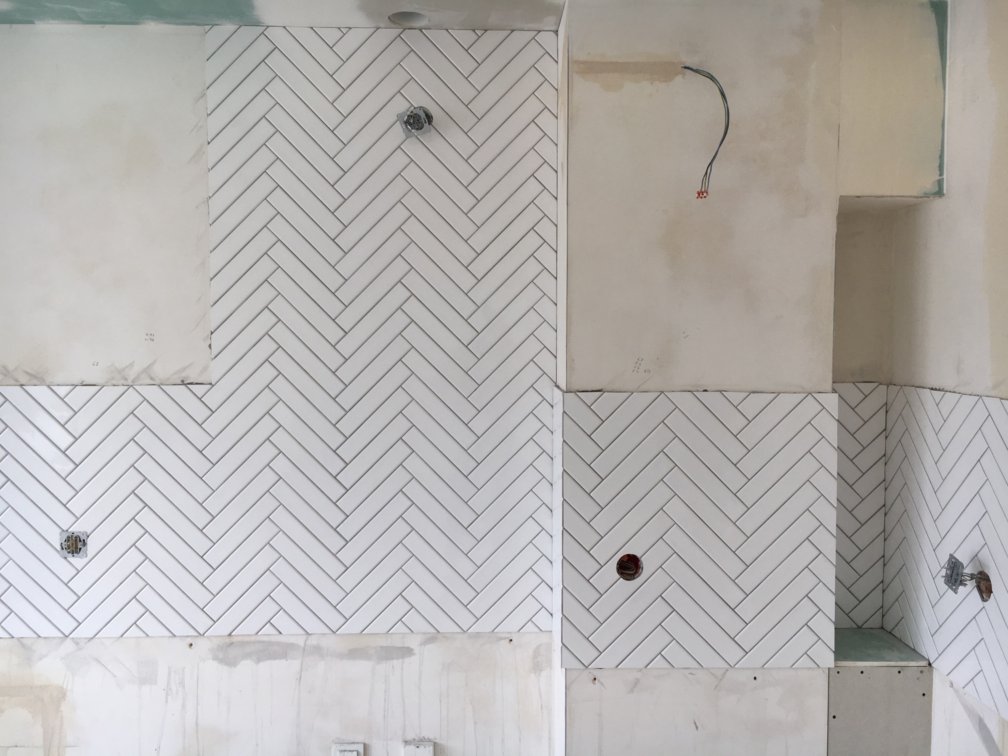 Białe, wąskie płytki ułożone w jodełkę na ścianie, widoczne puszki elektryczne i fragmenty nieotynkowanej ściany, etap prac wykończeniowych.