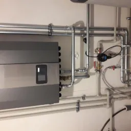 Szara pompa ciepła zamontowana na ścianie z licznymi rurami instalacyjnymi, zaworami i przewodami. Widoczne elementy instalacji grzewczej w pomieszczeniu technicznym.