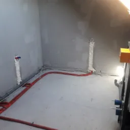 Surowy stan poddasza z instalacją hydrauliczną: czerwone rury ogrzewania podłogowego, szare rury kanalizacyjne i piony zabezpieczone pianką montażową przy ścianach, żółta skrzynka elektryczna w rogu.