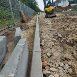 Montaż obrzeży betonowych na gruncie z gruzem, w tle żółta mini koparka i ogrodzenie panelowe.