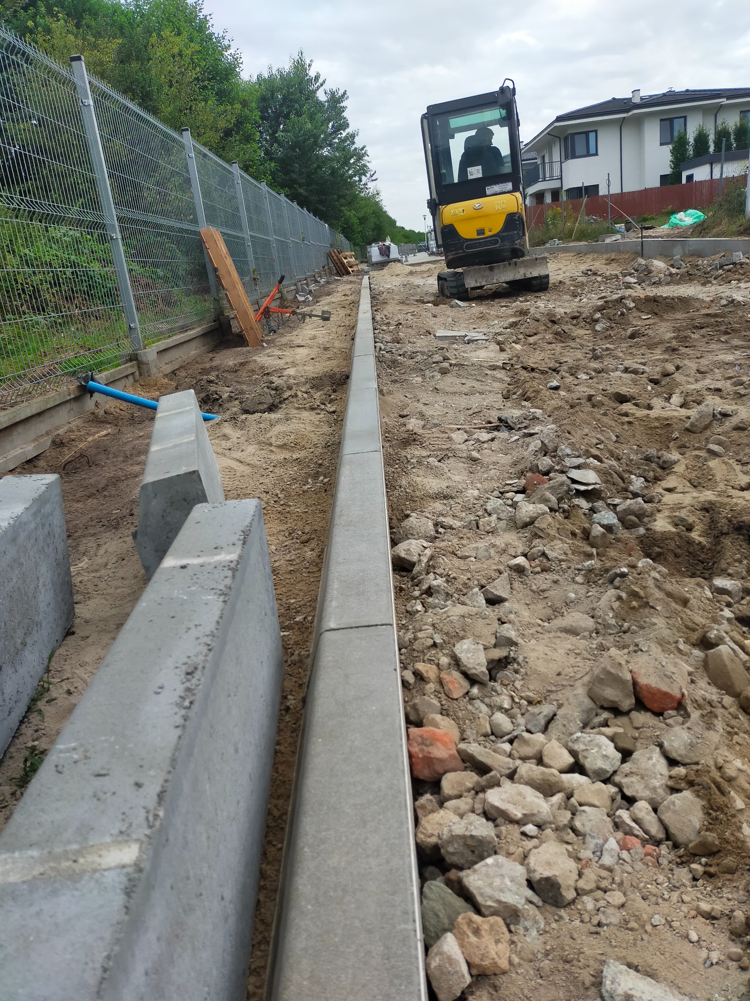 Montaż obrzeży betonowych na gruncie z gruzem, w tle żółta mini koparka i ogrodzenie panelowe.