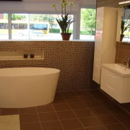 Nowoczesna łazienka z wolnostojącą białą wanną, brązowymi płytkami i szafką z umywalką. Za oknem widoczny żółty autobus.