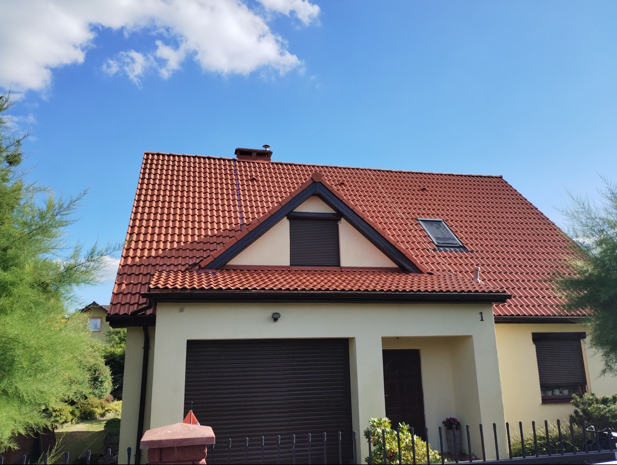 Dom jednorodzinny z czerwoną dachówką, widok z dołu na fasadę i dach. Na dachu okno dachowe i komin. Błękitne niebo z chmurami w tle. Z przodu ogrodzenie i zieleń.