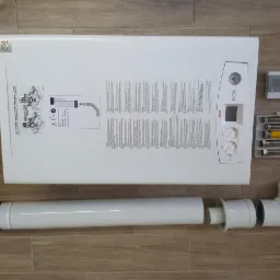 Nowy, biały gazowy kocioł kondensacyjny Unical KON z kompletnym systemem odprowadzania spalin i akcesoriami montażowymi, ułożony na podłodze imitującej drewno.