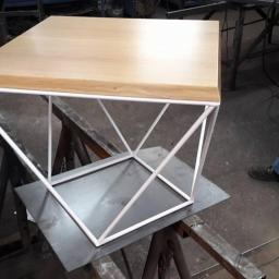 Szat-Com - Stolik kawowy z jasnego drewna z geometryczną, białą, metalową podstawą, umieszczony na metalowym arkuszu w warsztacie.