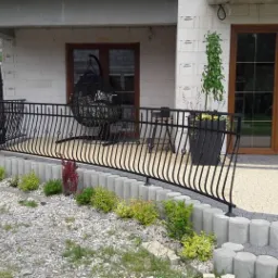 Czarna, gięta balustrada metalowa z ozdobnymi elementami na tarasie domu z białego bloczka, widoczny wiszący fotel, rośliny w donicach i częściowo zaparkowany samochód dostawczy.