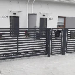 Nowoczesna, pozioma brama wjazdowa w kolorze antracytowym z furtką i skrzynkami na listy, na tle bliźniaczego domu z numerami 8B/1 i 8B/2.