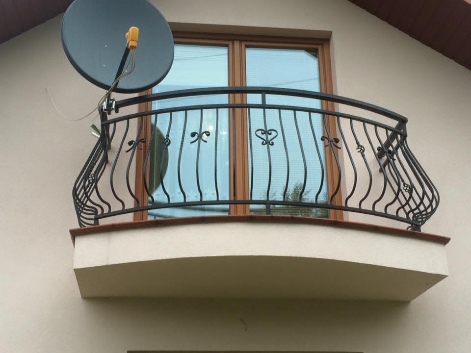 Czarna, ozdobna barierka balkonowa z motywami serc i kokardek, zamontowana na balkonie z brązową ramą okienną i anteną satelitarną w tle.