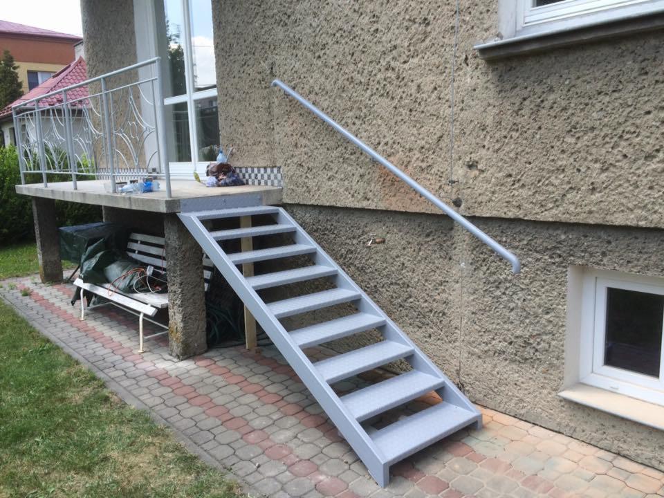 Zewnętrzne, metalowe schody z poręczą prowadzące na betonowy taras z ozdobną, metalową balustradą, widok z boku na tle chropowatej elewacji budynku.