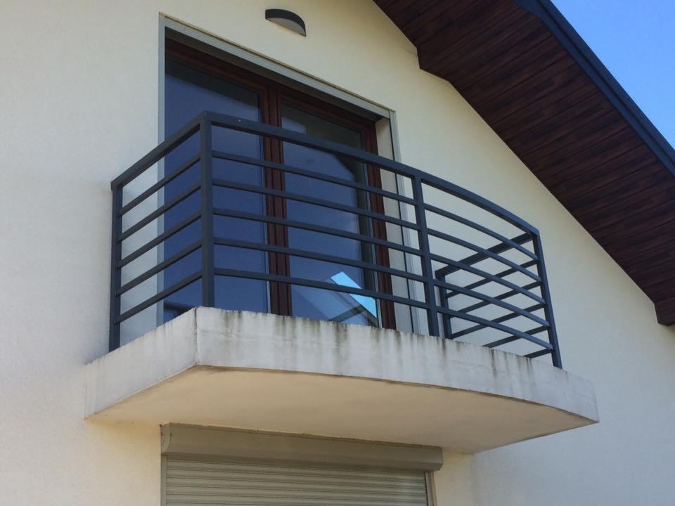 Czarna, metalowa balustrada balkonowa z poziomymi prętami i zaokrąglonym narożnikiem, zamontowana na betonowej płycie balkonu przy oknie z brązową ramą, na tle białej ściany budynku i fragmentu dachu.