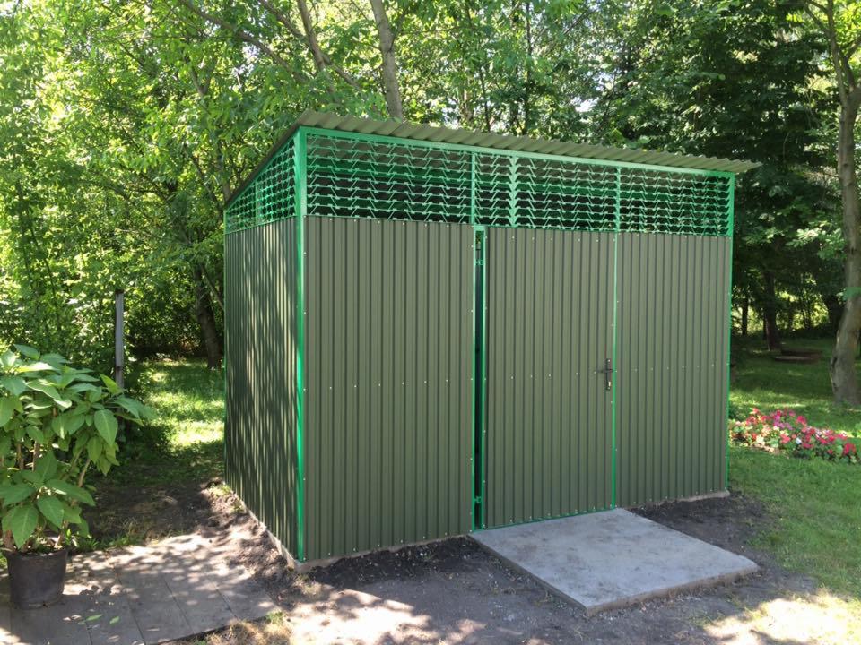 Zielony, blaszany garaż z dwuskrzydłowymi drzwiami, z ażurowym dachem, ustawiony na betonowej wylewce w ogrodzie. Widoczne drzewa i kwiaty w tle.