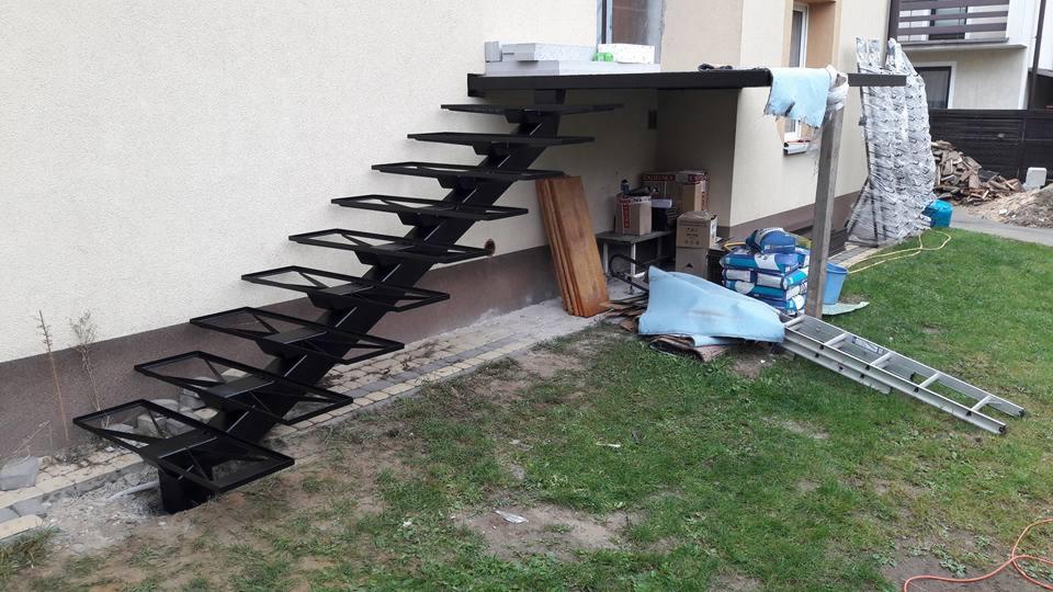 Czarne, metalowe schody zewnętrzne, o geometrycznym ażurowym wzorze stopni, przymocowane do jasnej ściany budynku, z widocznymi elementami budowlanymi i porządkiem na trawniku w tle.