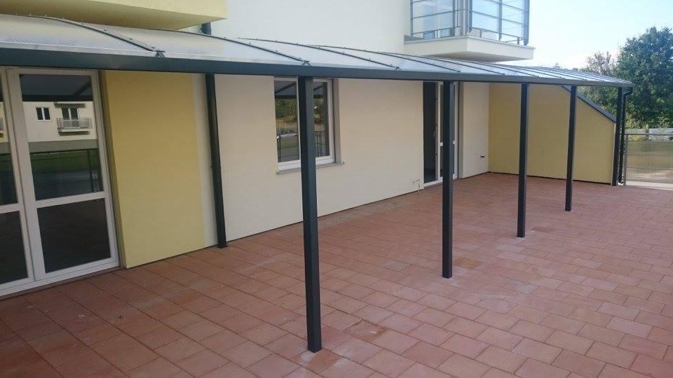 Zadaszenie tarasu z poliwęglanu na metalowej konstrukcji, wspartej na czterech słupach, przylegające do budynku z oknami i drzwiami na ceglanej posadzce.