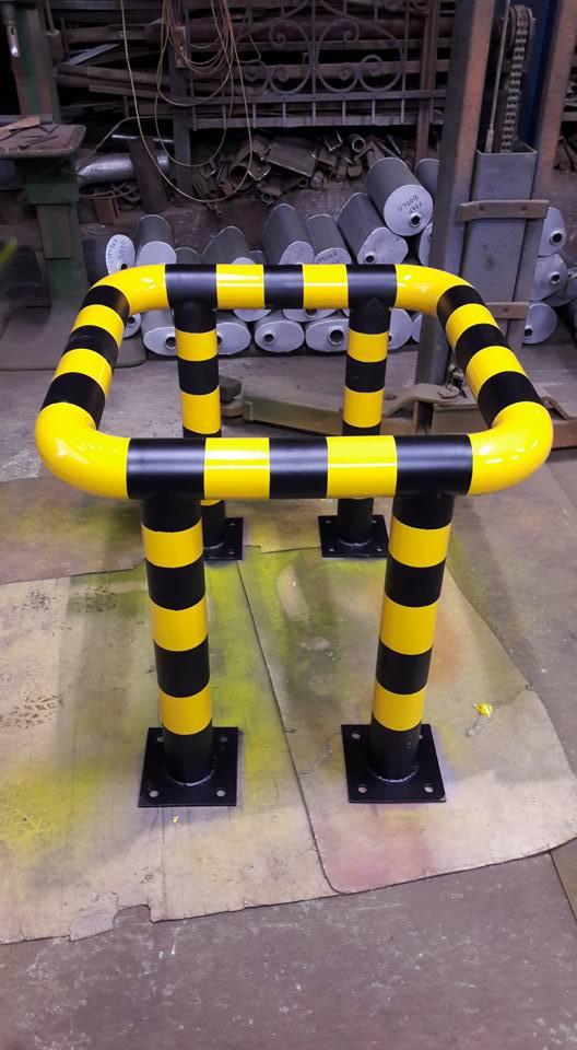 Metalowa bariera ochronna w czarno-żółte pasy, spawana do czarnych kwadratowych podstaw, w tle warsztat z metalowymi elementami.