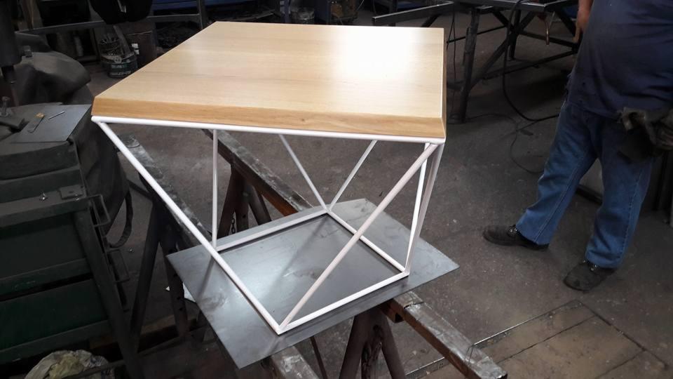 Stolik kawowy z jasnego drewna z geometryczną, białą, metalową podstawą, umieszczony na metalowym arkuszu w warsztacie.