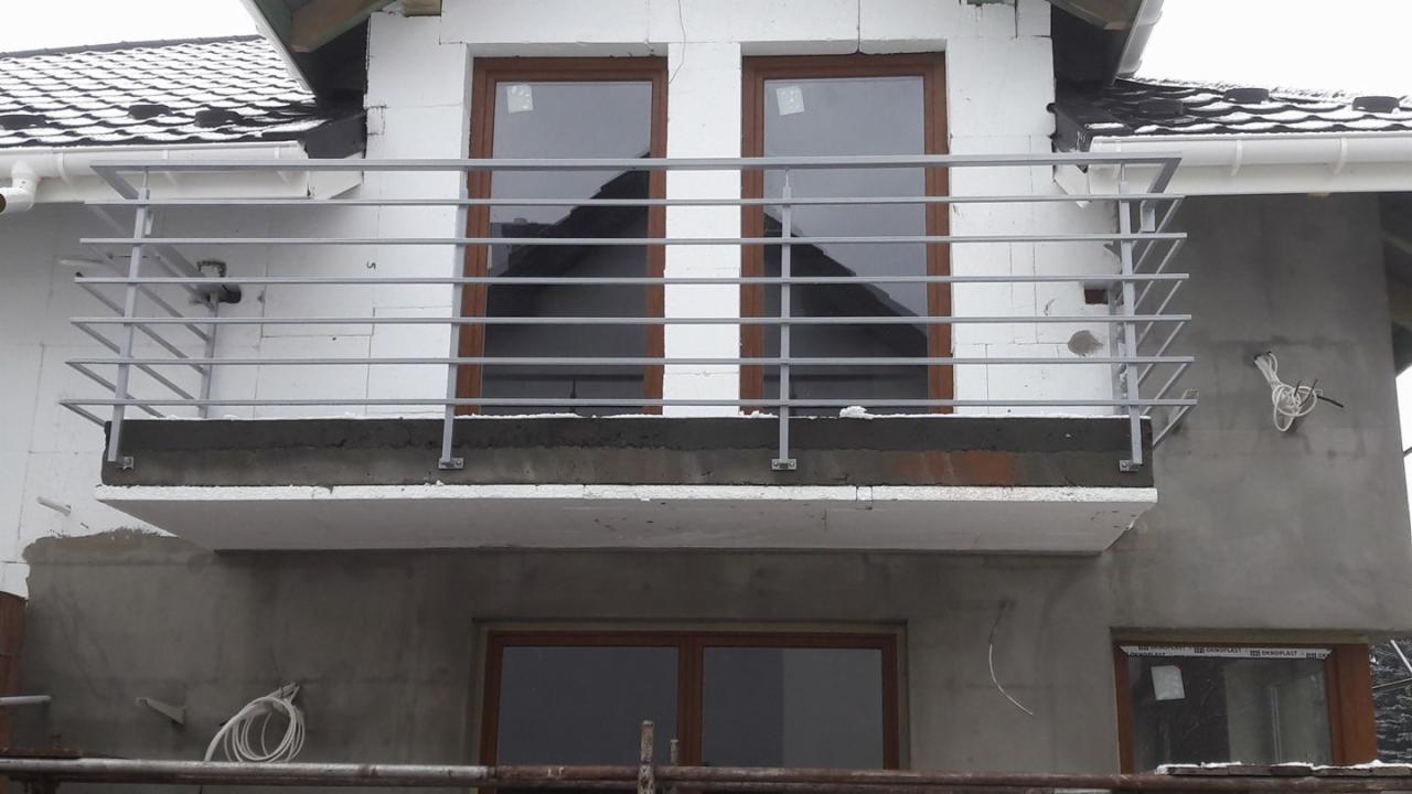 Surowy balkon z metalową barierką z poziomych prętów, widoczne niedokończone prace wykończeniowe elewacji i wystające kable elektryczne.