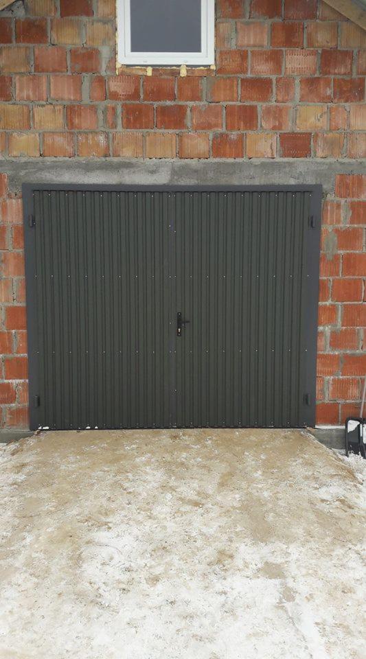 Zamknięta, szara brama garażowa z blachy falistej, zamontowana w surowej, ceglanej ścianie budynku z małym oknem u góry, przed bramą śnieg z błotem.