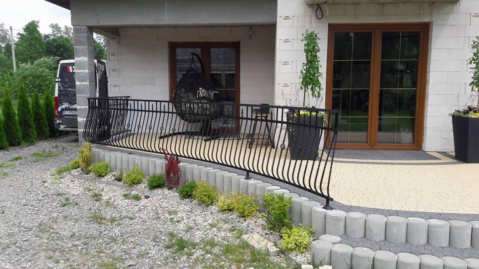 Czarna, gięta balustrada metalowa z ozdobnymi elementami na tarasie domu z białego bloczka, widoczny wiszący fotel, rośliny w donicach i częściowo zaparkowany samochód dostawczy.