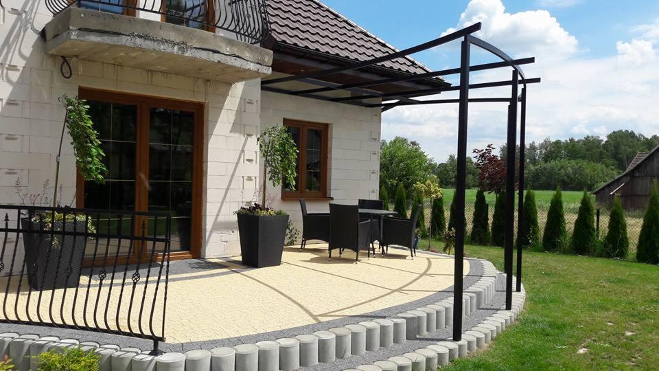 Zadaszony taras z czarną, metalową konstrukcją, wyłożony jasną kostką brukową, z ciemnymi meblami ogrodowymi i ozdobnymi roślinami w donicach, na tle domu z białego pustaka i zielonego ogrodu.