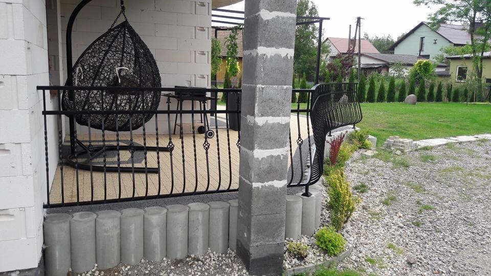 Zadaszony taras z czarną, metalową balustradą z dekoracyjnymi elementami, betonowymi słupkami i wiszącym fotelem w kształcie łzy.