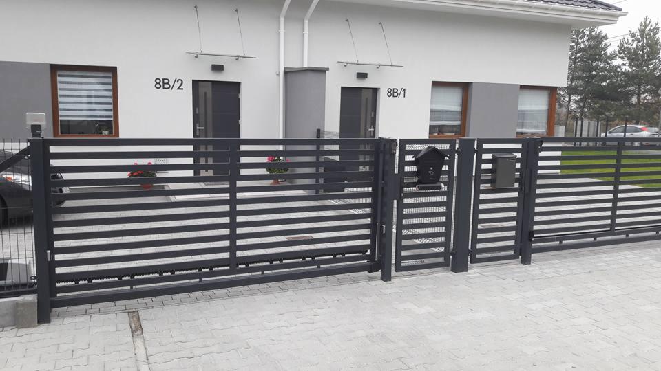 Nowoczesna, pozioma brama wjazdowa w kolorze antracytowym z furtką i skrzynkami na listy, na tle bliźniaczego domu z numerami 8B/1 i 8B/2.