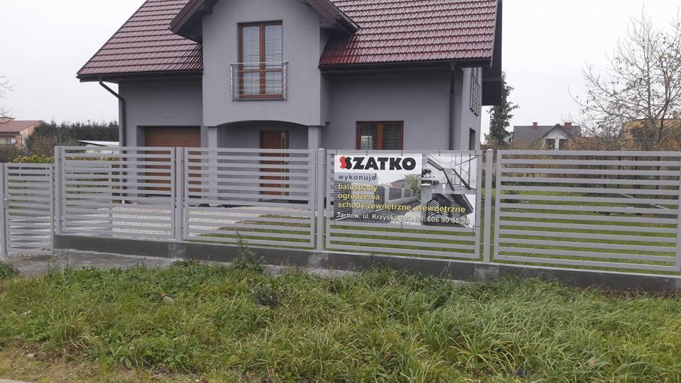 Nowoczesne ogrodzenie posesji z poziomych, szarych profili metalowych, z przesuwną bramą wjazdową i furtką, na tle domu z szarą elewacją i brązowym dachem, z widoczną reklamą firmy na ogrodzeniu.
