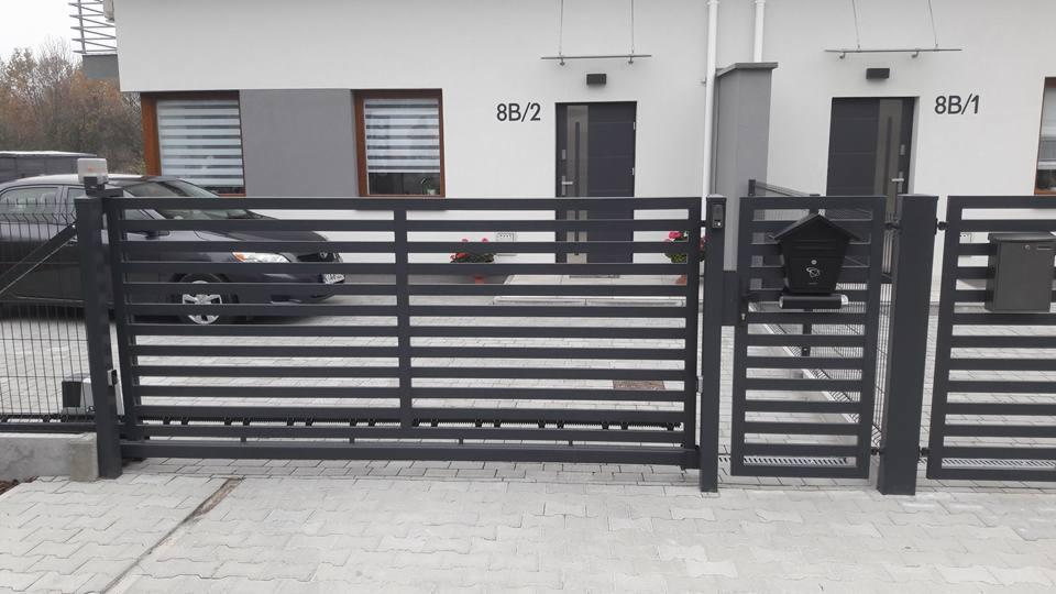 Nowoczesna, pozioma, metalowa brama wjazdowa w kolorze grafitowym, zautomatyzowana, z widocznym napędem, na tle bliźniaczych domów z numerami 8B/1 i 8B/2 oraz zaparkowanym samochodem.