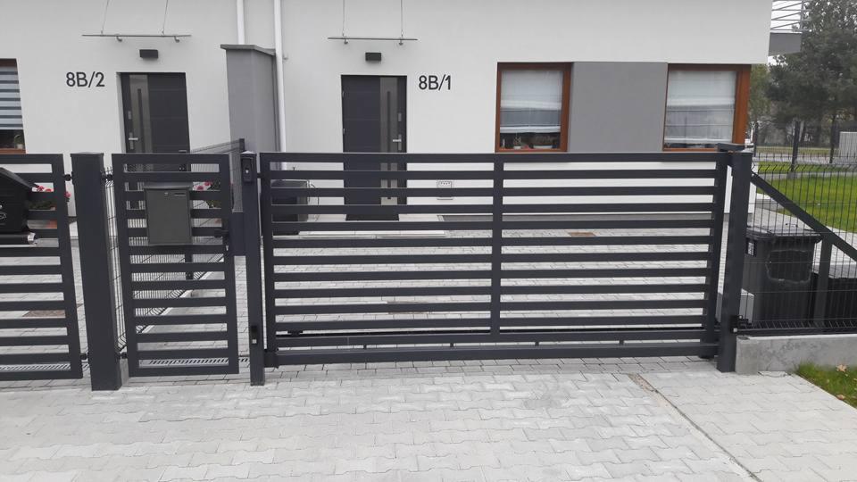 Nowoczesna, przesuwna brama wjazdowa z poziomymi profilami, w kolorze antracytowym, przed bliźniaczym domem z numerami 8B/1 i 8B/2.