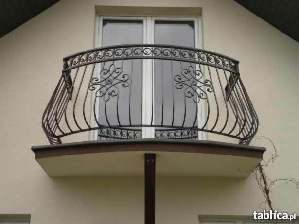 Kuty balkon z czarną, zdobioną balustradą na tle białej ściany budynku i okna.