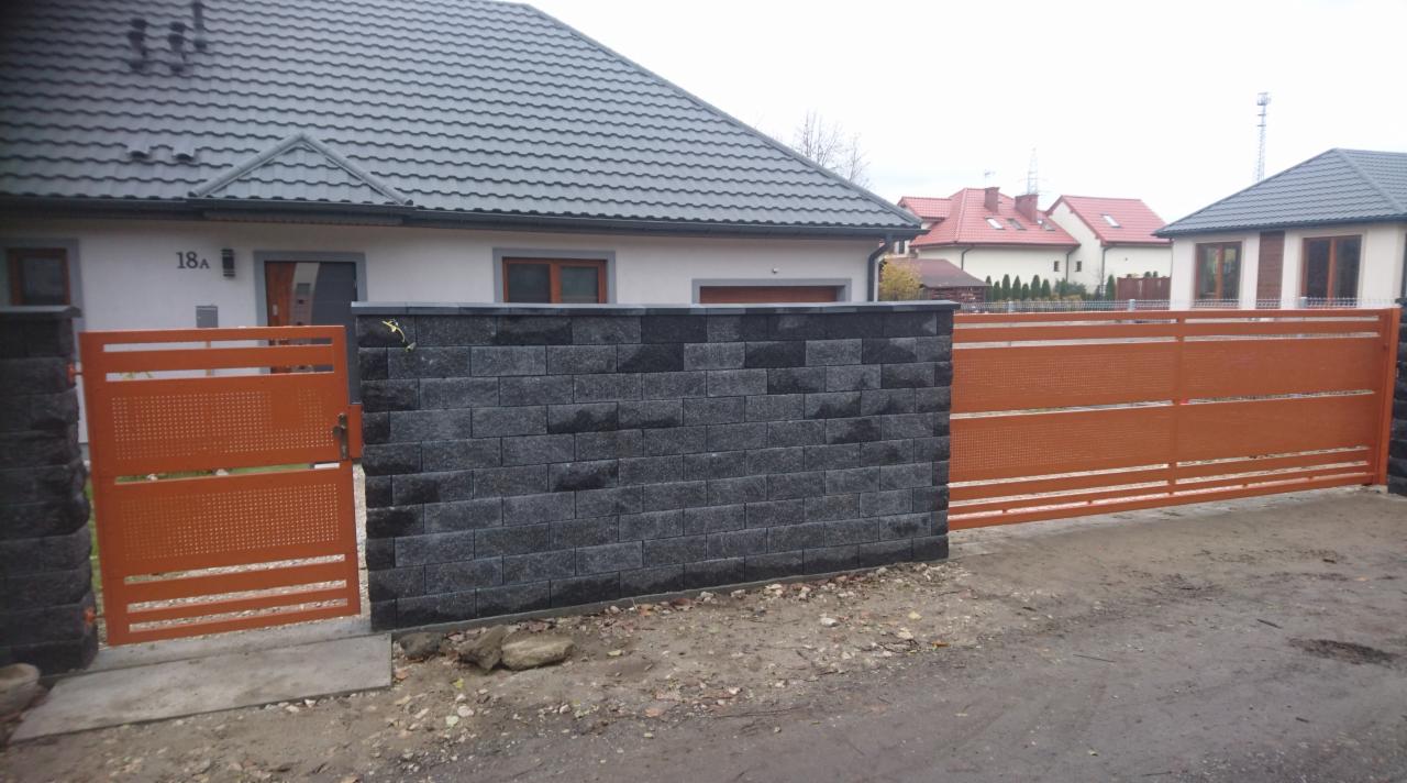 Nowoczesna posesja z przesuwną bramą w kolorze ceglastym i furtką o podobnym designie, uzupełniona murem z ciemnych, nieregularnych bloczków.