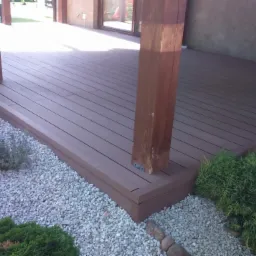 Brunatny taras kompozytowy z drewnianymi słupami, otoczony jasnym żwirem i zielenią, przylegający do budynku z ciemnym tynkiem.