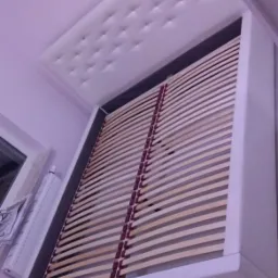 Białe łóżko z tapicerowanym zagłówkiem w trakcie montażu, widoczne drewniane listwy stelaża i fragment ściany z grzejnikiem.