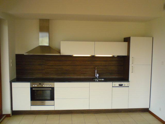 Nowoczesna kuchnia z białymi szafkami, ciemnym blatem i brązowym panelem ściennym, oświetlona listwą LED pod szafkami wiszącymi, widoczny srebrny okap i sprzęt AGD.